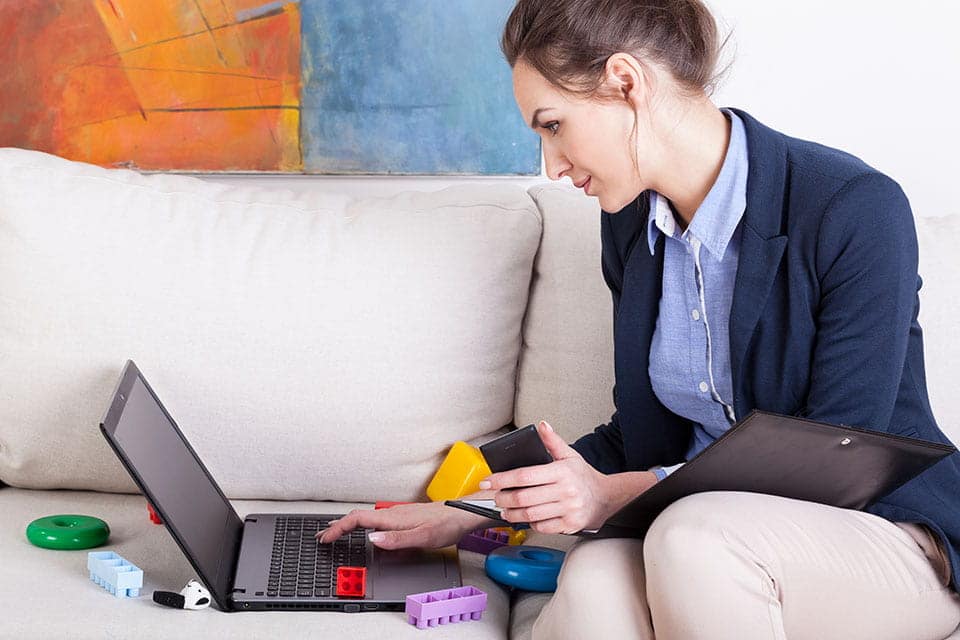 Is This Work at Home Paycheck Legitimate? employee sitting on a couch with a calculator and laptop with childrens toys