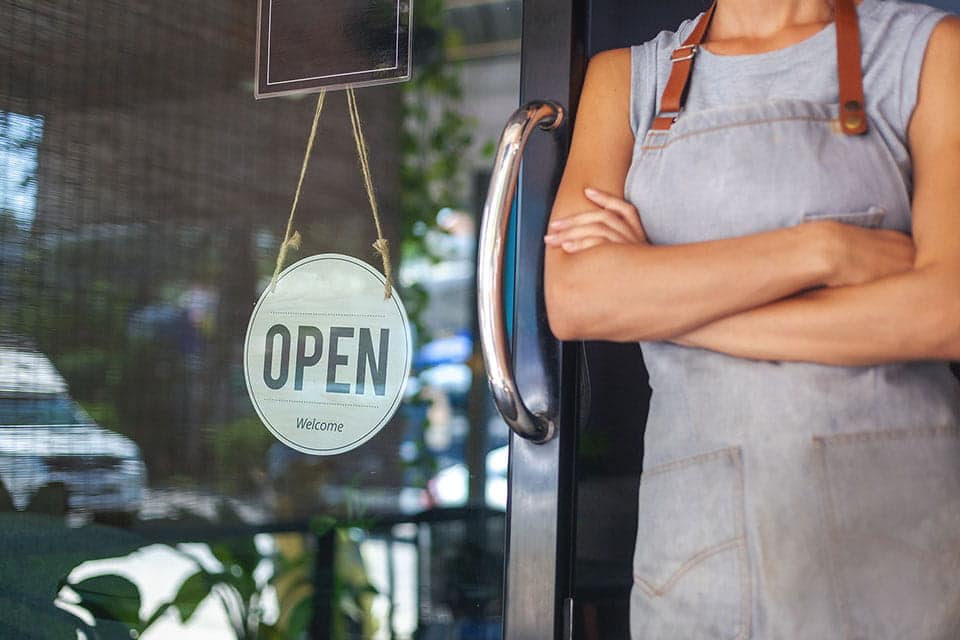 Do’s and Don’ts of Small Business Payroll employee with apron standing infront of a door with an open welcome sign